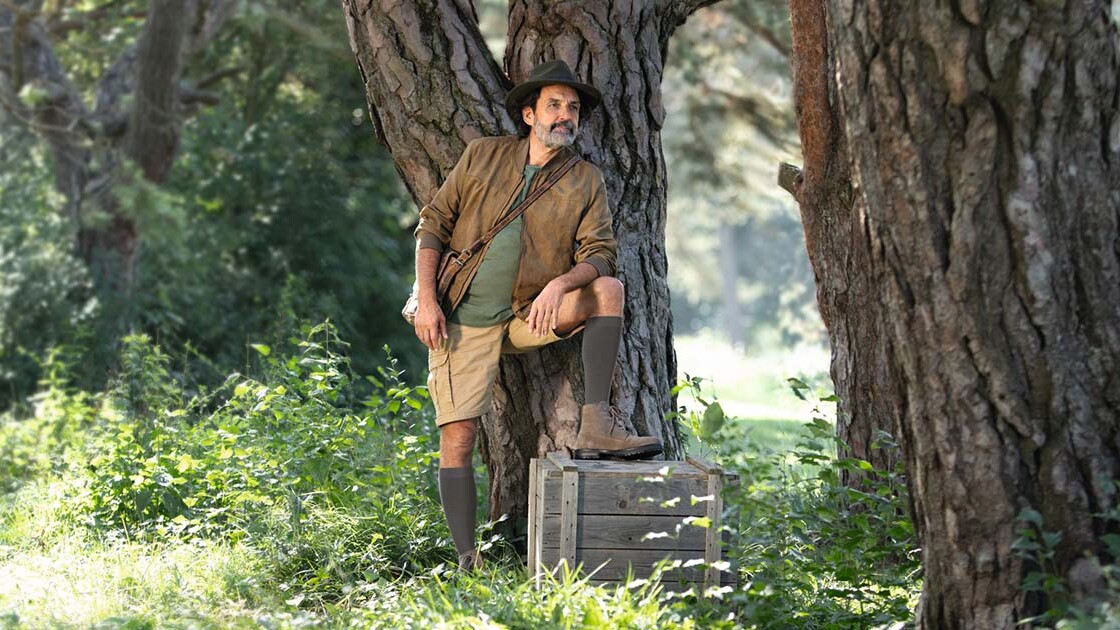 Ein Mann mit Entdecker-Outfit steht in der Natur und schaut in die Ferne. Er trägt kniehohe Kompressionsstrümpfe. Ein Mann mit Entdecker-Outfit steht in der Natur und schaut in die Ferne. Er trägt kniehohe Kompressionsstrümpfe.