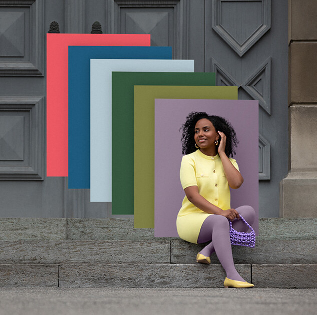 Juzo Trend Colours Batik A woman sits on stone steps in front of a gate. She wears a light yellow two-piece outfit with a purple compression garment. Behind her, you can see colour fields in the trend colours.