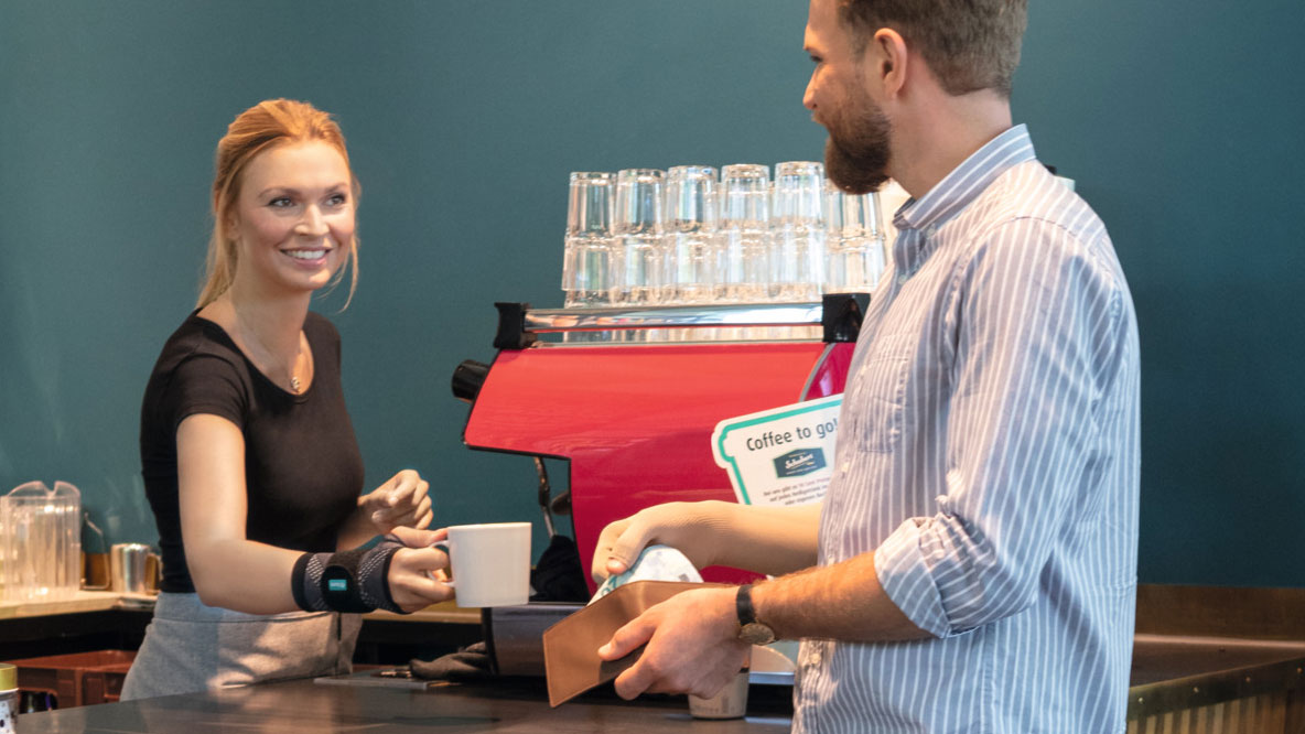 Kellnerin trägt Handgelenkbandage JuzoFlex Manu Xtra und reicht Kunden eine Tasse über den Thresen Kellnerin trägt Handgelenkbandage JuzoFlex Manu Xtra und reicht Kunden eine Tasse über den Thresen