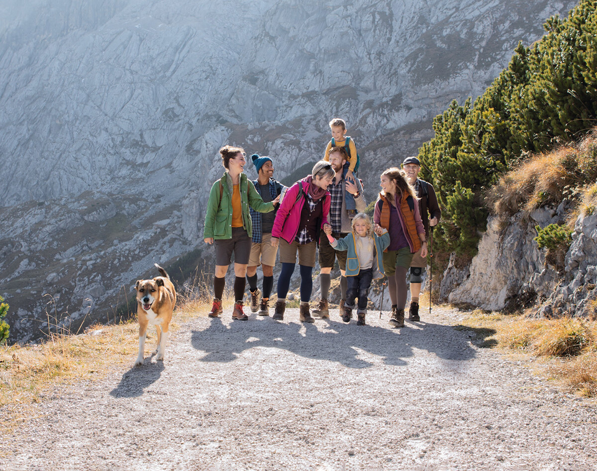 Groupe de randonneurs sur un sentier de montagne, certains portant des bas de contention et des bandages, accompagnés d'un chien devant un décor rocheux.