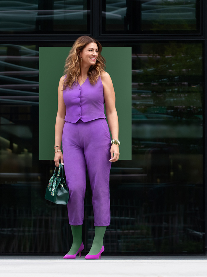 A smiling woman in a purple two-piece outfit stands in front of a building wearing a Strong Green compression garment